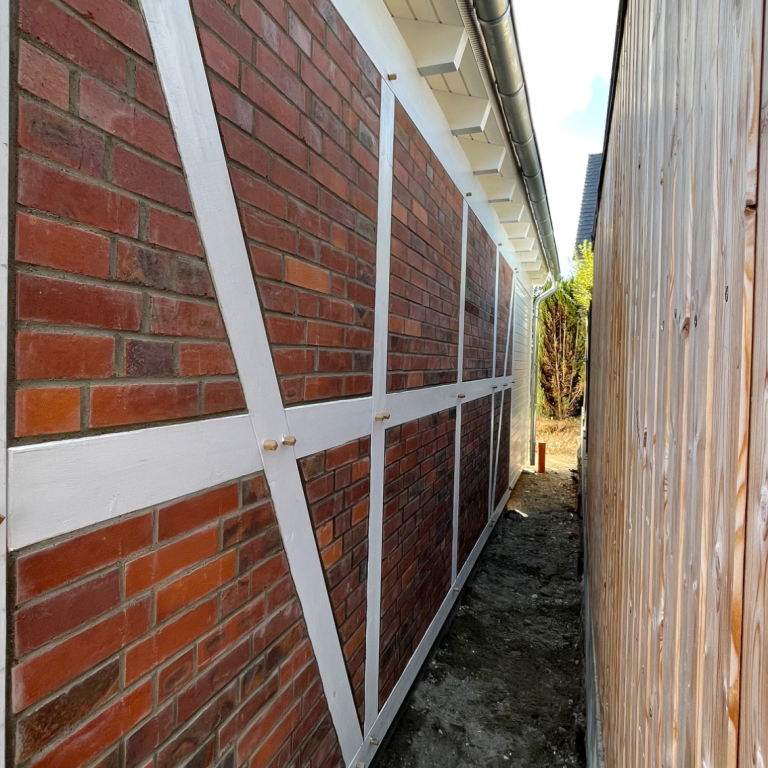 Fachwerkartige Verstrebungen zur Stabilisierung einer Backsteinwand im Außenbereich, ausgeführt in einem engen Durchgang.