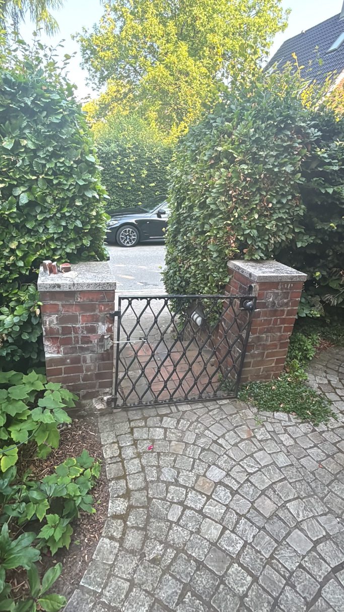 Eingangsbereich mit zwei verwitterten Klinkersäulen und altem Metalltor; Vorgarten wirkt offen und baulich undefiniert vor der Sanierung
